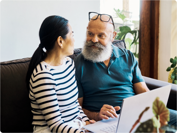 Older couple sitting on a couch at home, looking at each other and smiling while using a laptop, with indoor plants in the background.