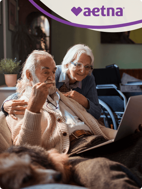 Senior couple on the couch looking at a computer together with an off-white and purple swoop in the top right corner framing the Aetna logo