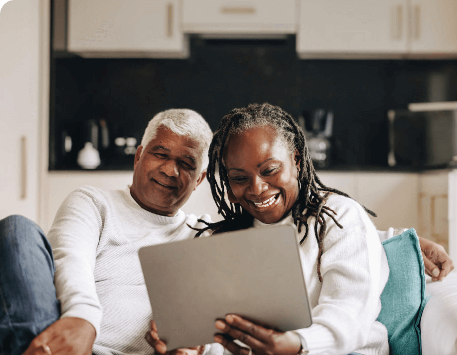 A smiling couple sits on a couch, looking at a tablet together. The man has gray hair, and the woman has long braided hair, both in white sweaters.