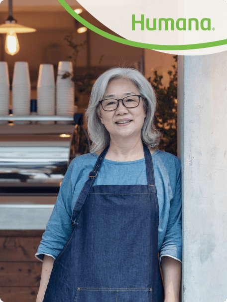 Smiling older woman with gray hair and glasses, wearing a denim apron, standing outside a cozy café. Humana logo appears in the corner.