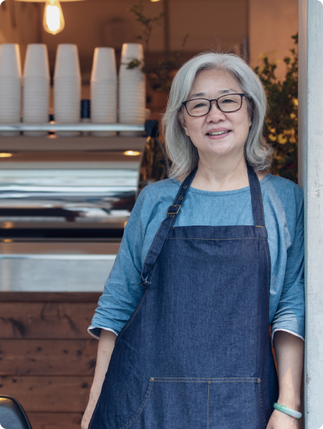A woman with gray hair and glasses, wearing a denim apron, stands smiling in front of a coffee shop counter with white coffee cups stacked on top.