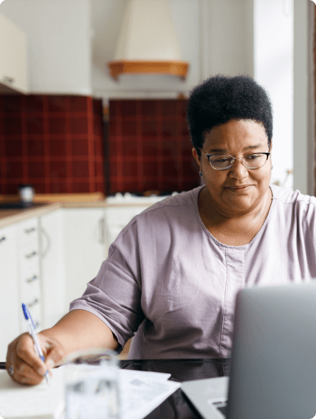 A woman sitting at the kitchen table filling out paper work and looking at her laptop.