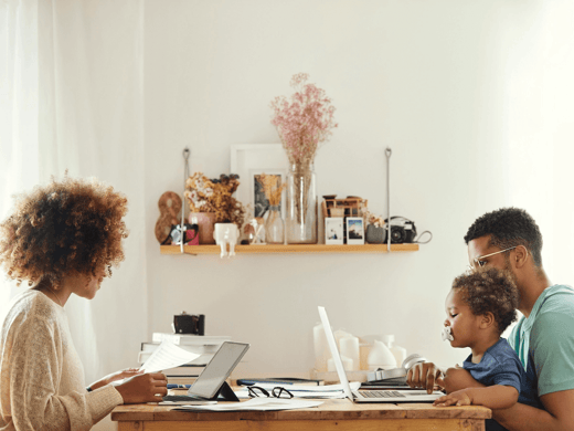 A mother and father at the dining table with laptops, while the father is holding his child in his lap.
