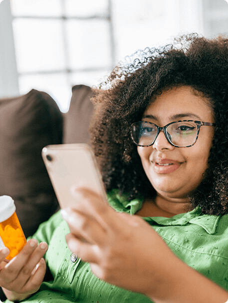 A young woman in a green shirt holds a prescription bottle and uses a smartphone while sitting comfortably on a couch. A young woman in a green shirt holds a prescription bottle and uses a smartphone while sitting comfortably on a couch.