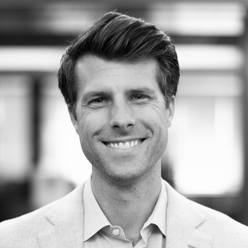 Black-and-white headshot of Jesse Waldron, Chief Financial Officer, smiling and wearing a light-colored suit jacket, with a blurred background.